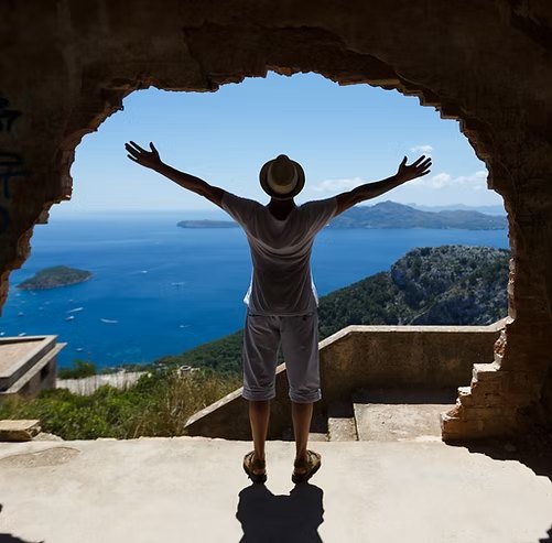 A woman with arms raised looks out over the sea through a ruined stone arch.